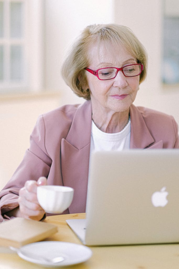 Aged female in eyeglasses with cup checking important documents on netbook on blurred background